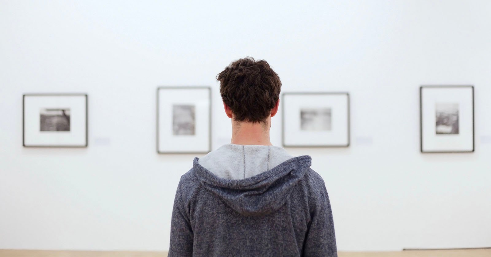 A man with curly hair, seen from behind, wearing a gray hoodie, viewing a series of framed black and white photographs on a white wall in an art gallery.